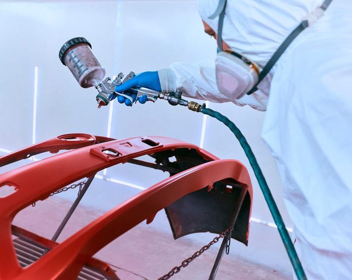 Un hombre está pintando con aerosol el parachoques de un coche rojo.