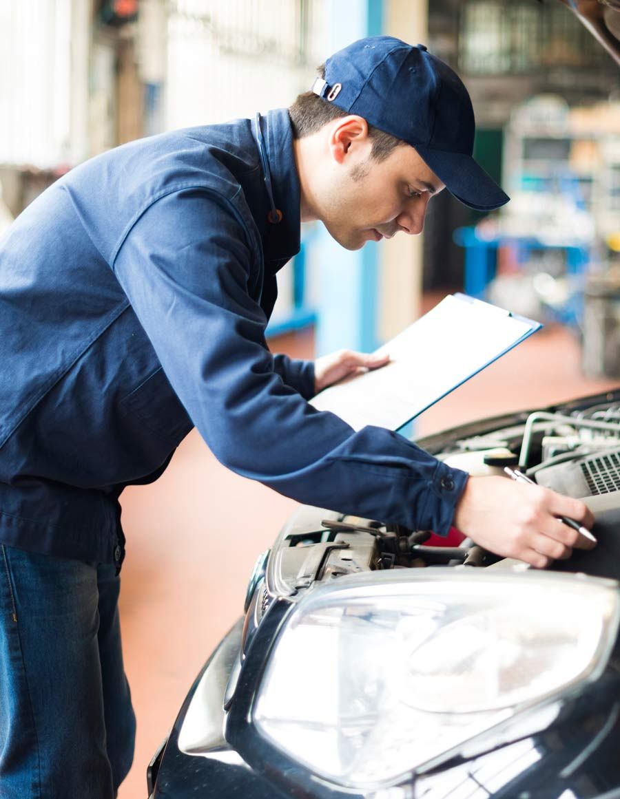 Un hombre mira debajo del capó de un automóvil mientras sostiene un portapapeles.