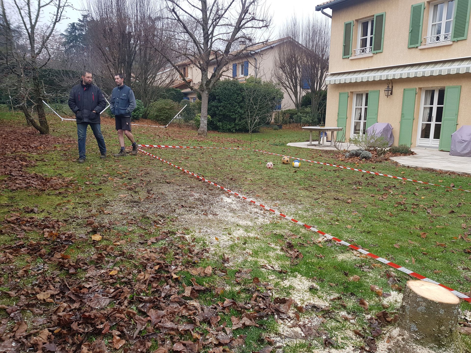 Deux personnes se tiennent debout sur une pelouse à côté d'une maison, une ligne de ruban adhésif rouge et blanc barrant le sol.
