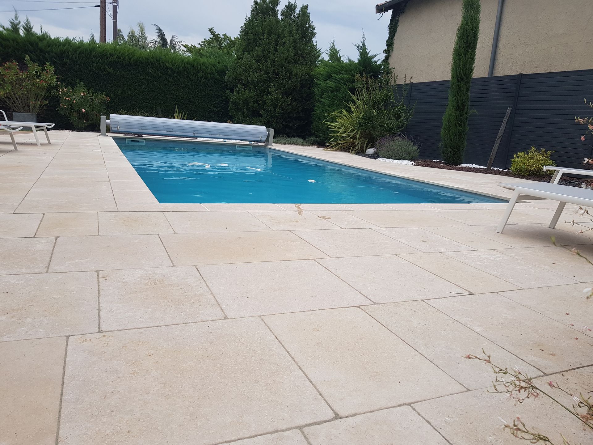 Piscine dans le jardin avec terrasse en pierre claire, eau bleue et chaise longue blanche près d'une clôture bordée de haies.