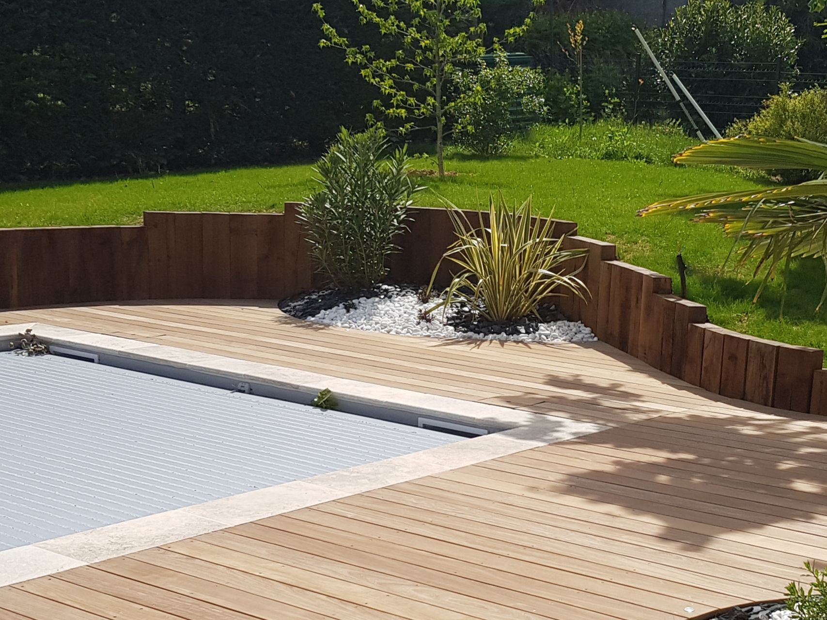 Terrasse de piscine dans le jardin avec planches de bois clair, jardinières en bois et pelouse verte ensoleillée