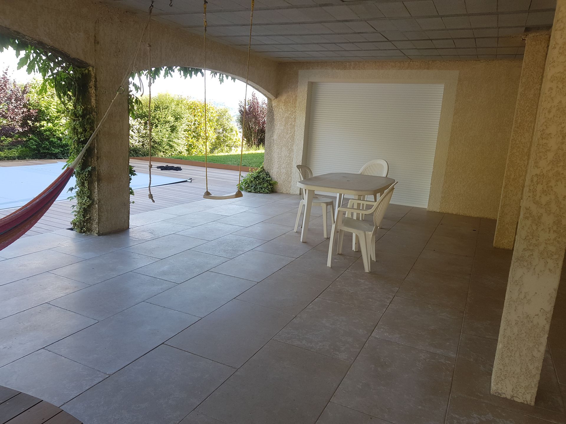 Terrasse couverte avec sol carrelé, table et chaises blanches, et un hamac donnant sur la verdure et l'eau.