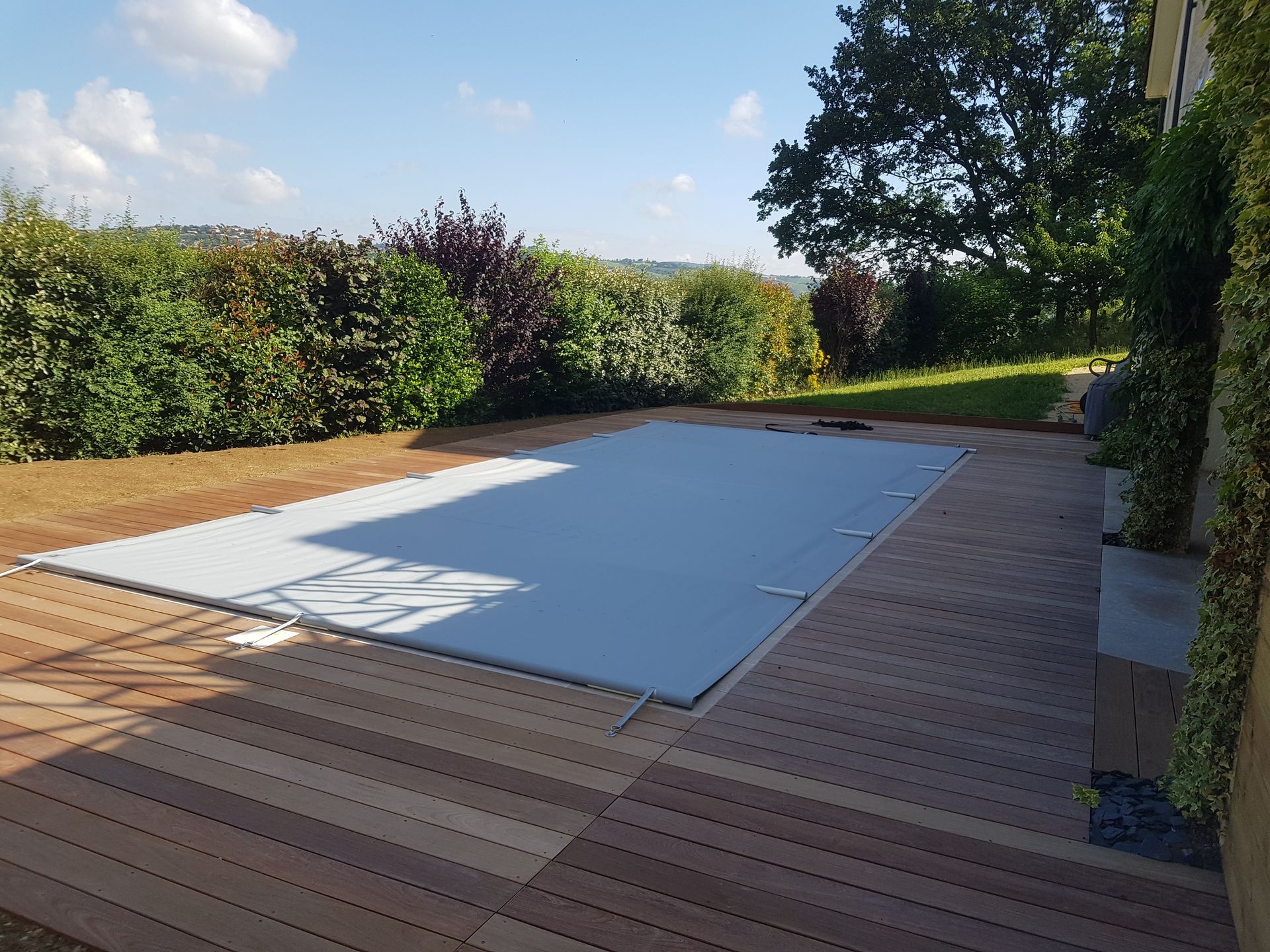 Terrasse en bois dans le jardin avec piscine rectangulaire couverte, bordée de haies et d'arbres.