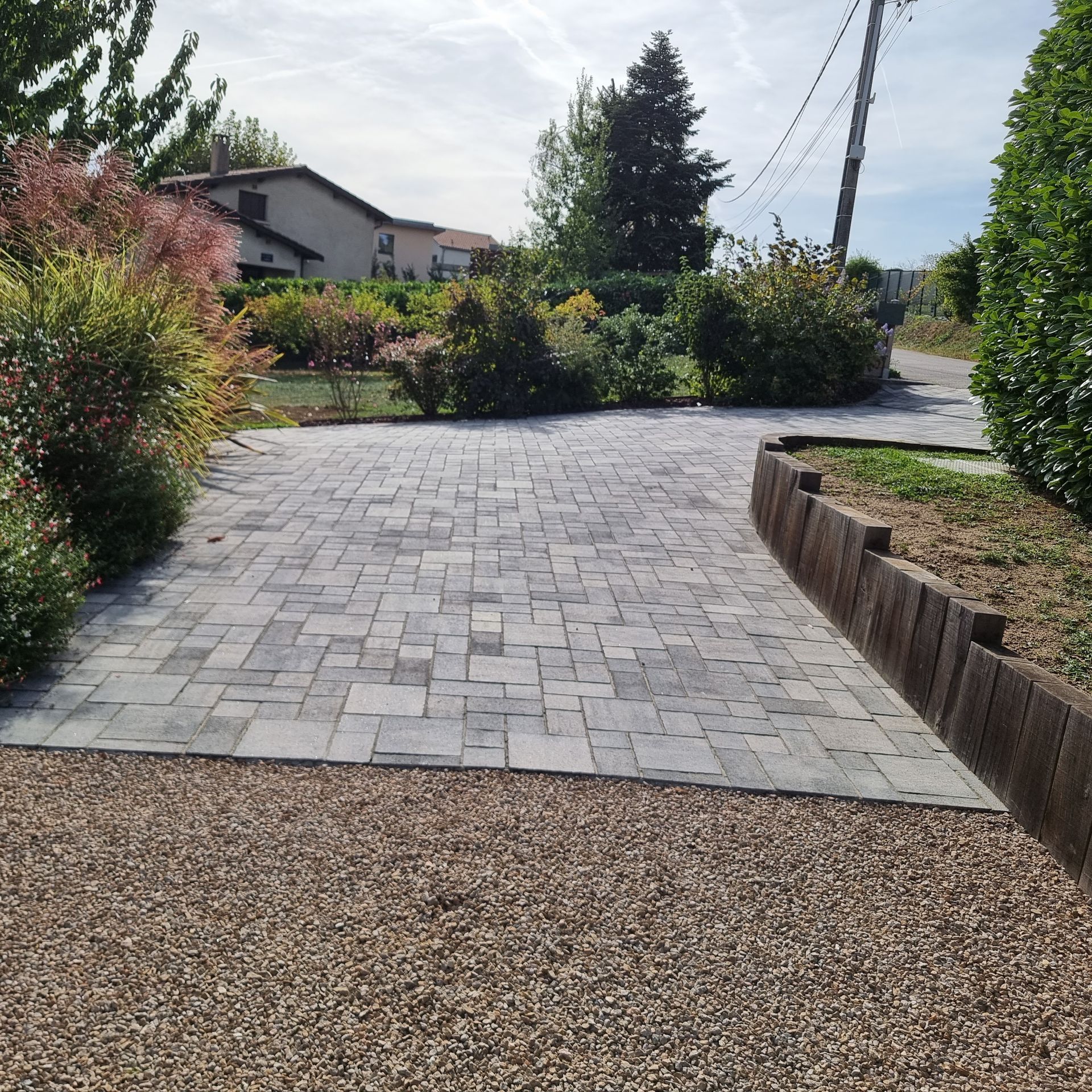 Terrasse pavée dans la cour arrière avec bordure de gravier, jardinière surélevée et arbustes sous un ciel ensoleillé.