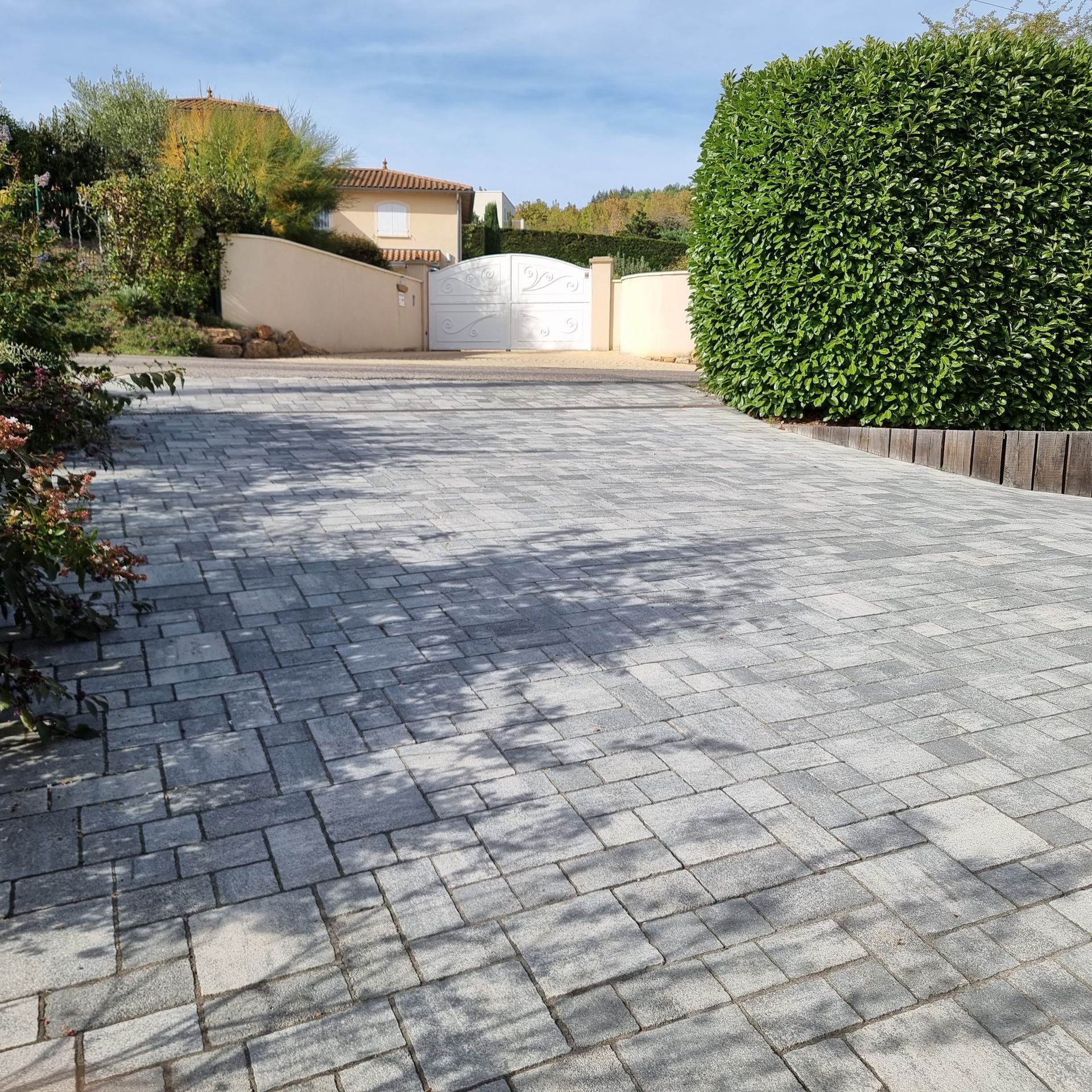 Allée pavée de dalles grises menant à un portail blanc, bordée de haies et de plantes de jardin.
