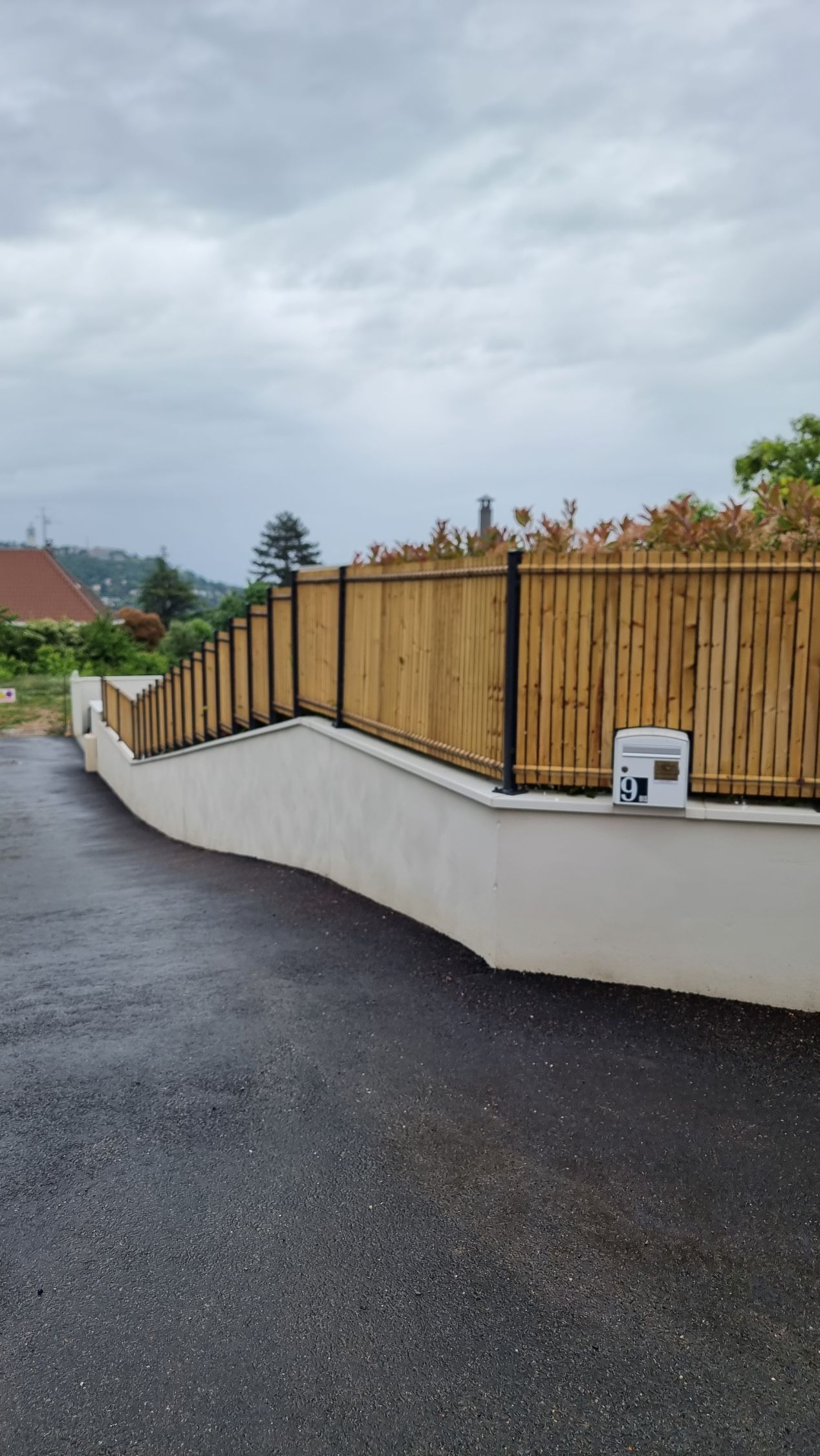 Allée mouillée longeant un mur blanc et une clôture en bambou sous un ciel nuageux