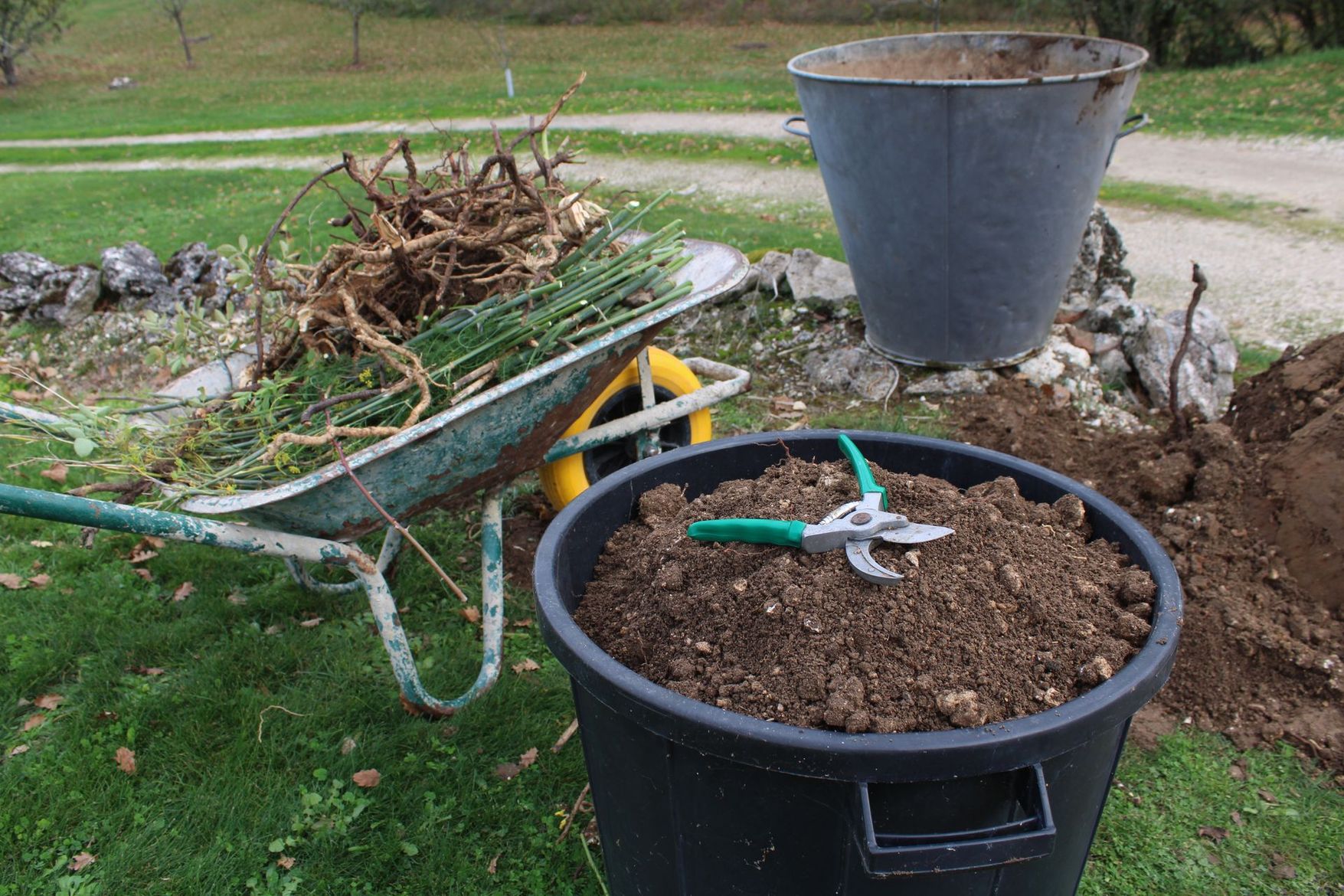 Des outils de nettoyage de jardin dans une cour, avec une brouette de branches et un grand bac à compost noir.