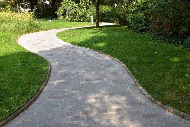 Allée en pierre incurvée traversant un parc verdoyant bordé d'arbres et de pelouse.
