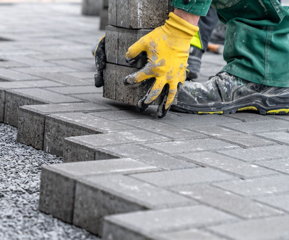 Un ouvrier portant des gants jaunes et un pantalon vert pose des pavés gris sur un trottoir.