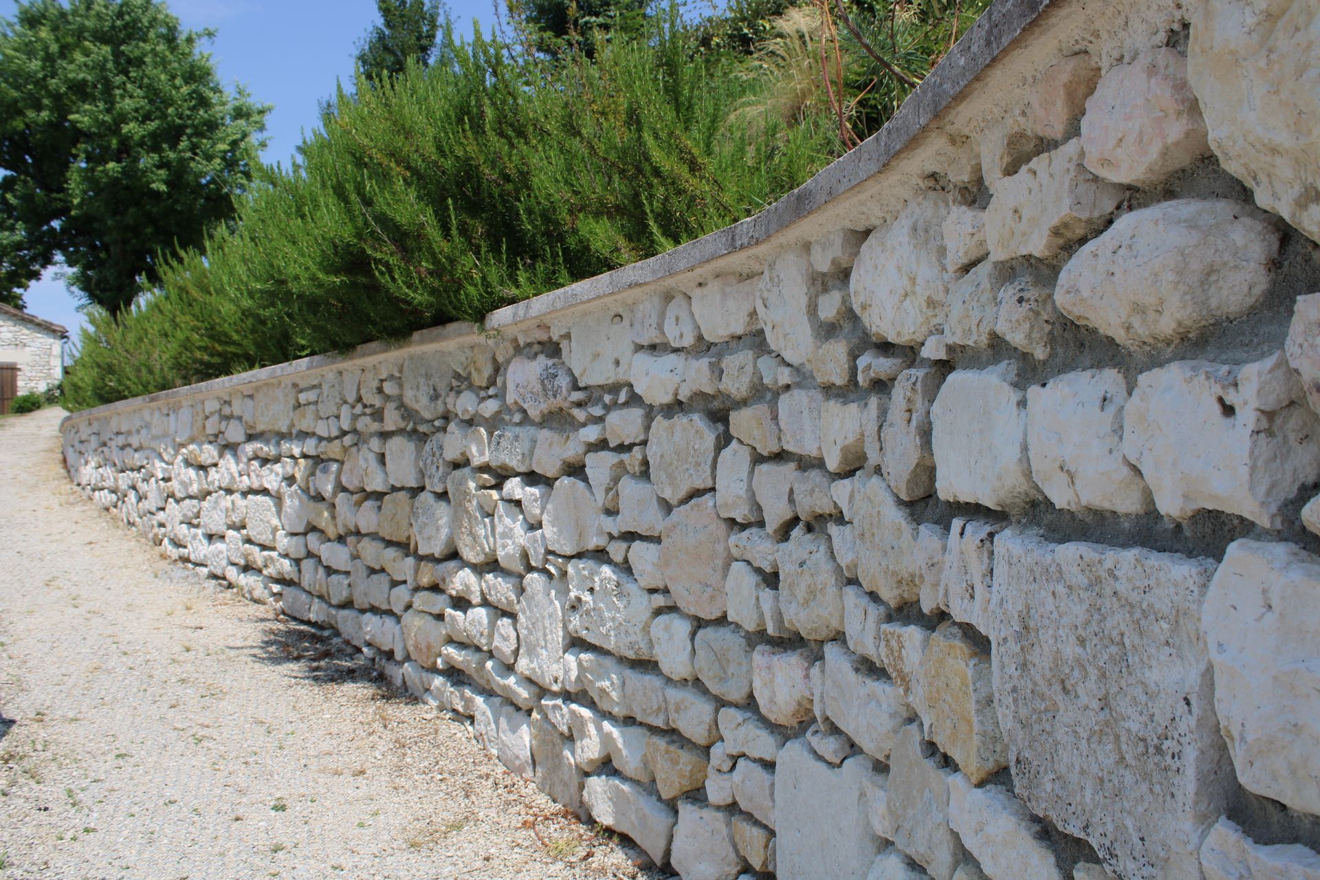 Mur de soutènement en pierre serpentant le long d'un chemin de gravier bordé d'arbustes et d'arbres verdoyants.
