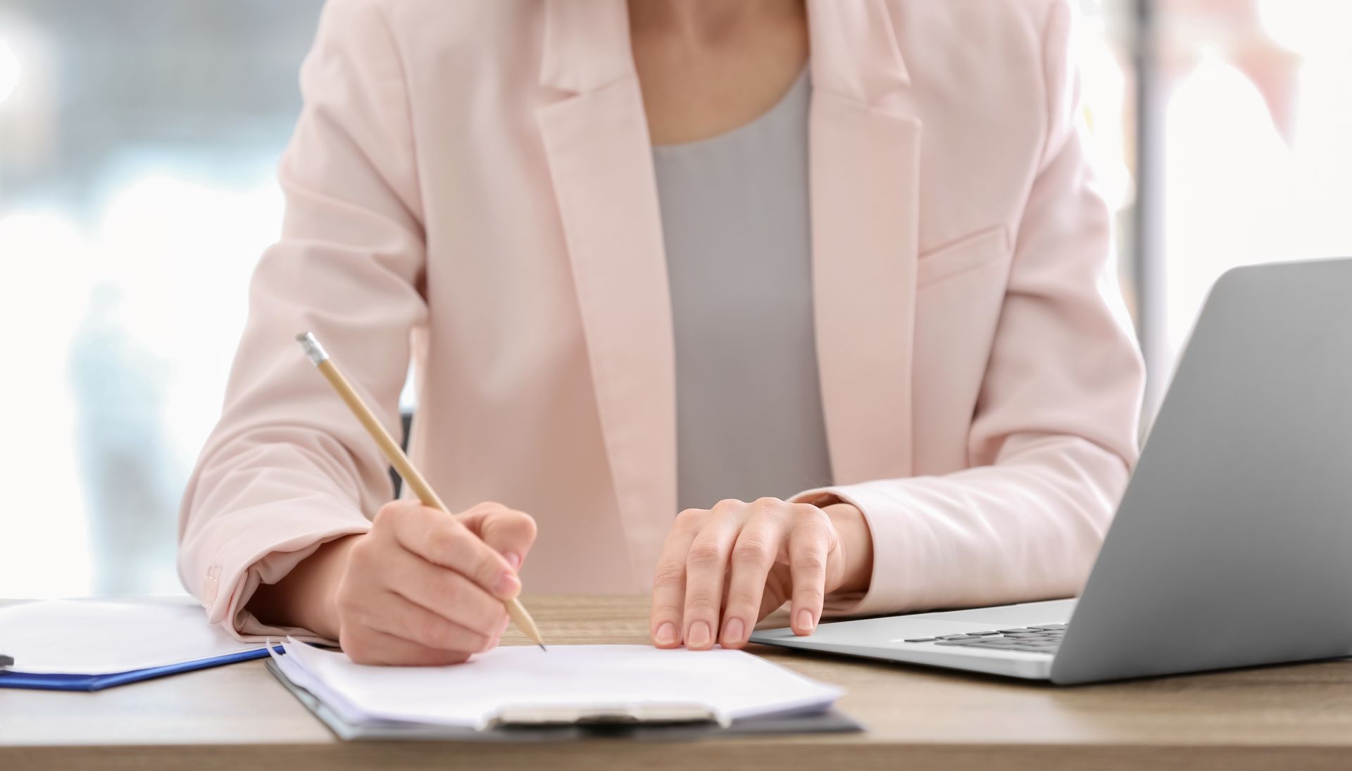 Mujer con chaqueta rosa escribiendo en un escritorio con una computadora portátil.