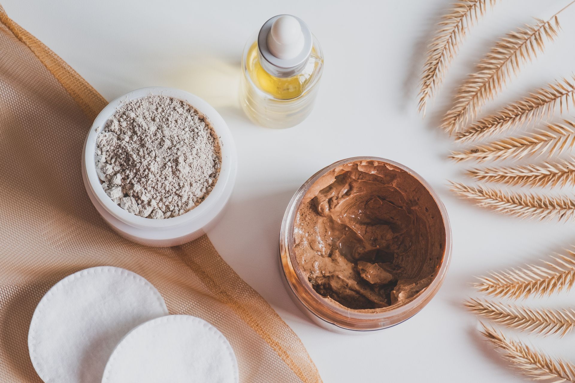 Produits de soin disposés sur une surface blanche : poudre d’argile, masque pour le visage, flacon d’huile, disques de coton et tiges de blé.