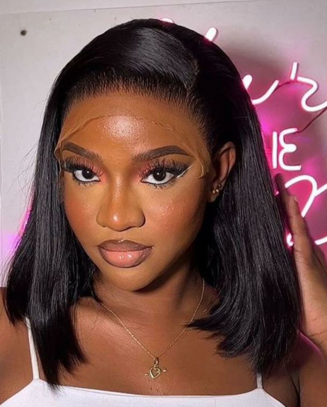 Une femme aux cheveux noirs coupés au carré regarde l'objectif. Elle porte une perruque à l'implantation naturelle et est maquillée ; la lumière est celle d'une enseigne lumineuse rose.
