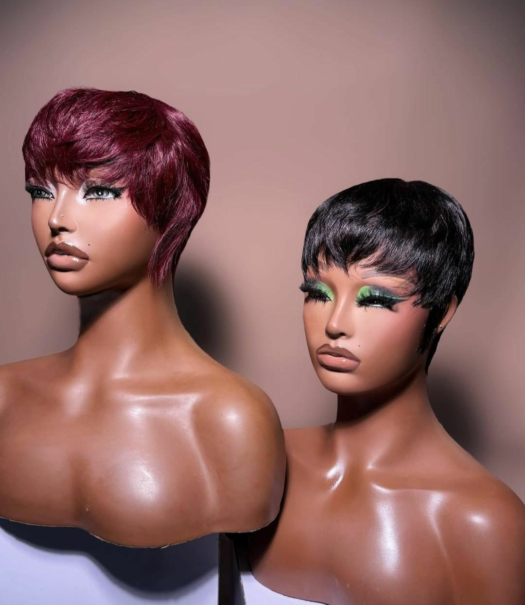 Deux têtes de mannequin avec des perruques courtes : l’une bordeaux, l’autre noire. Toutes deux sont fortement maquillées et ont le teint hâlé.