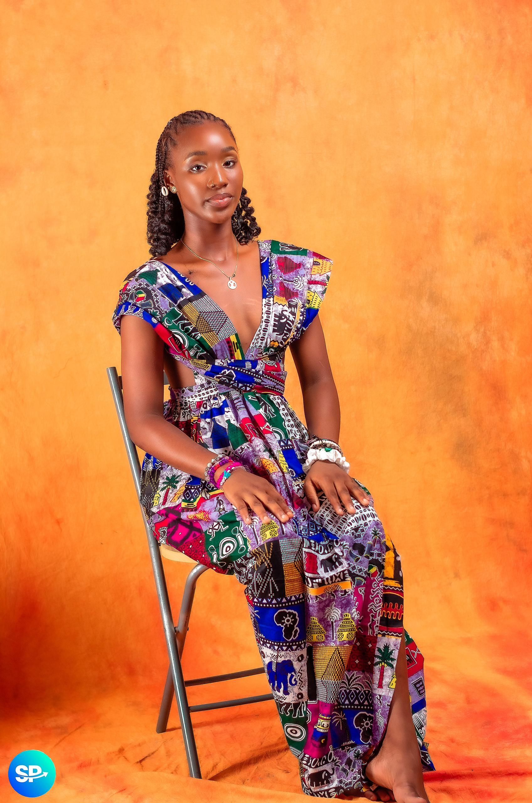Femme vêtue d'une robe à motifs colorés, assise sur une chaise devant un fond orange.
