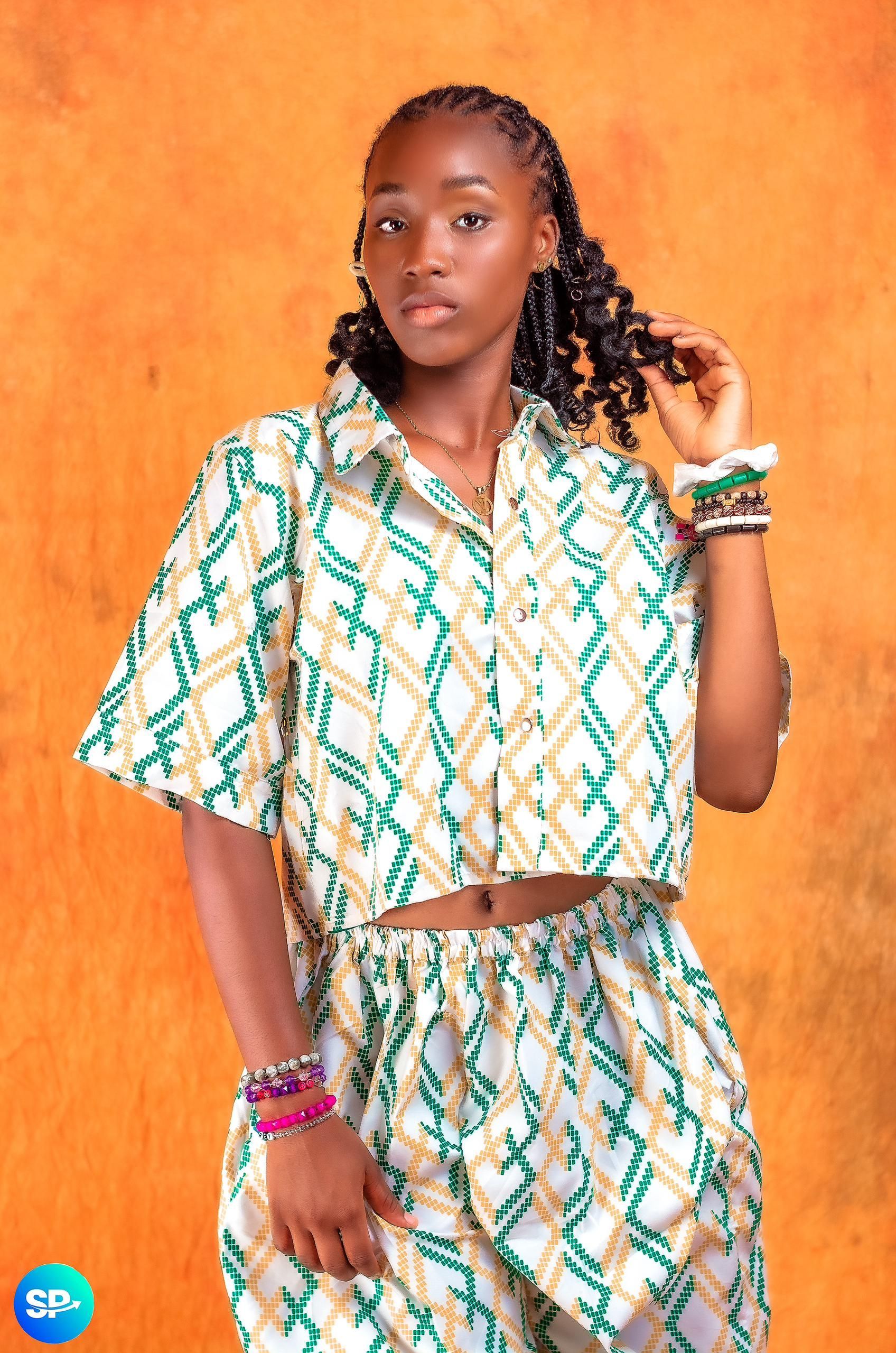 Femme vêtue d'un ensemble crop top et pantalon à motifs verts et blancs, tenant ses cheveux ; fond orange.