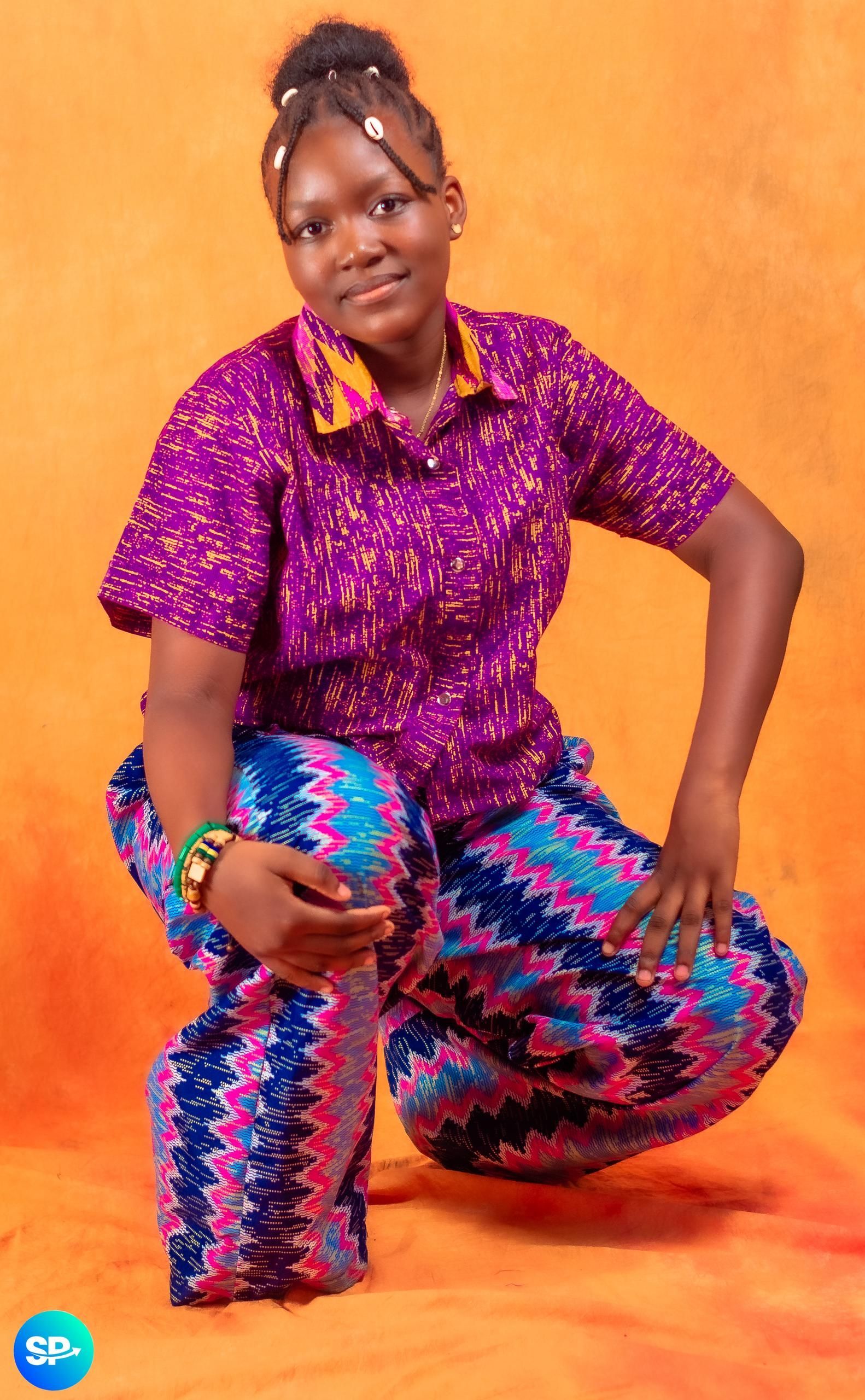 Femme vêtue d'une tenue à motifs violets et bleus, accroupie, souriant sur un fond orange.