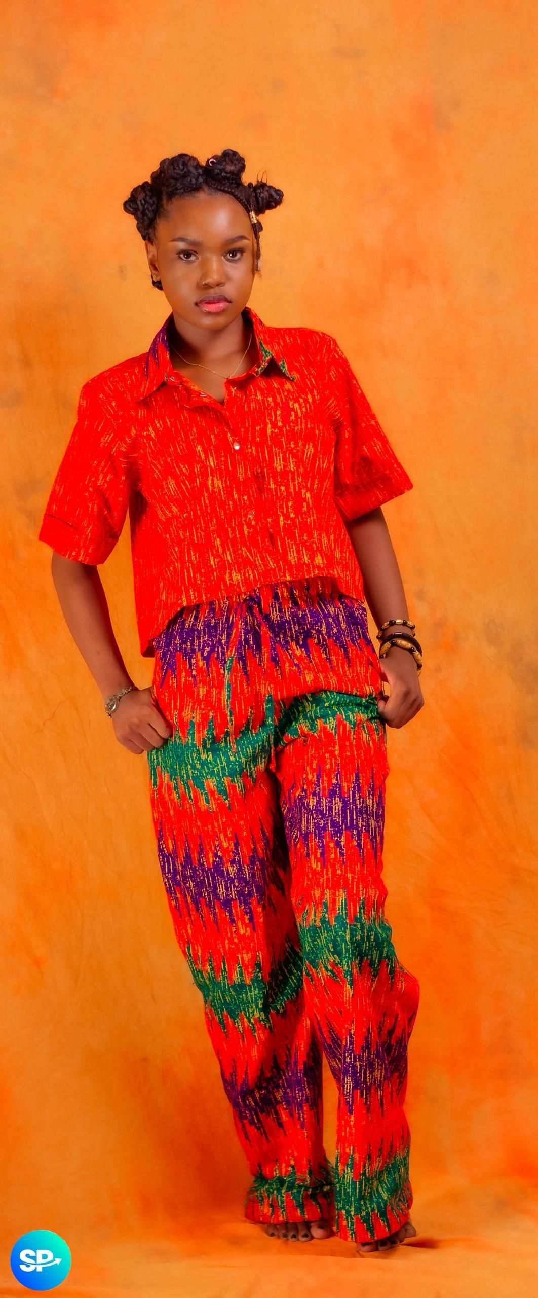 Femme en chemise et pantalon rouges à motifs, les mains dans les poches, posant sur fond orange.