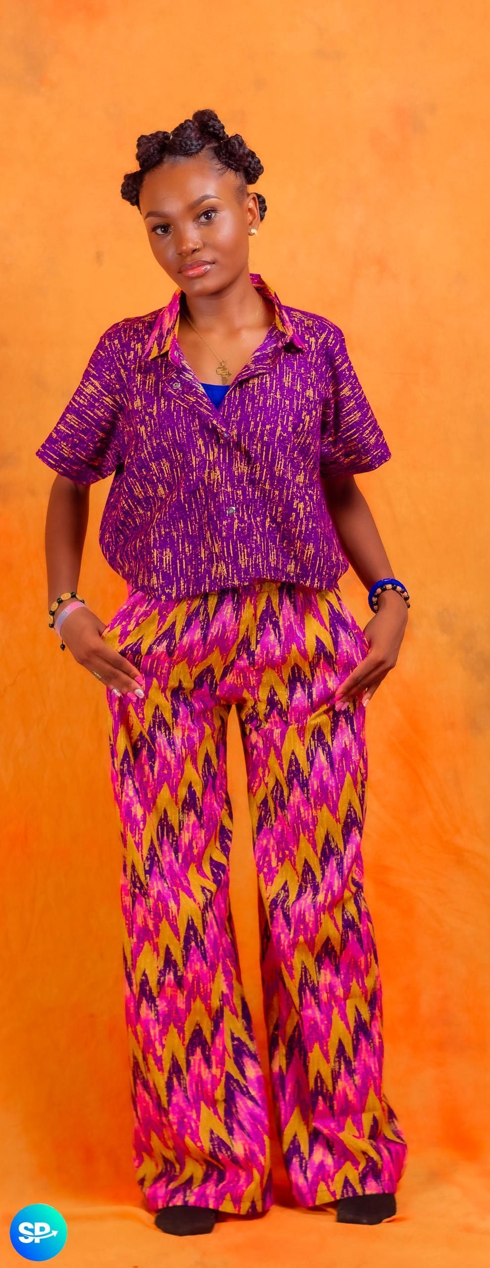 Une personne vêtue d'une tenue violette à motifs se tient debout, les mains dans les poches, sur un fond orange.