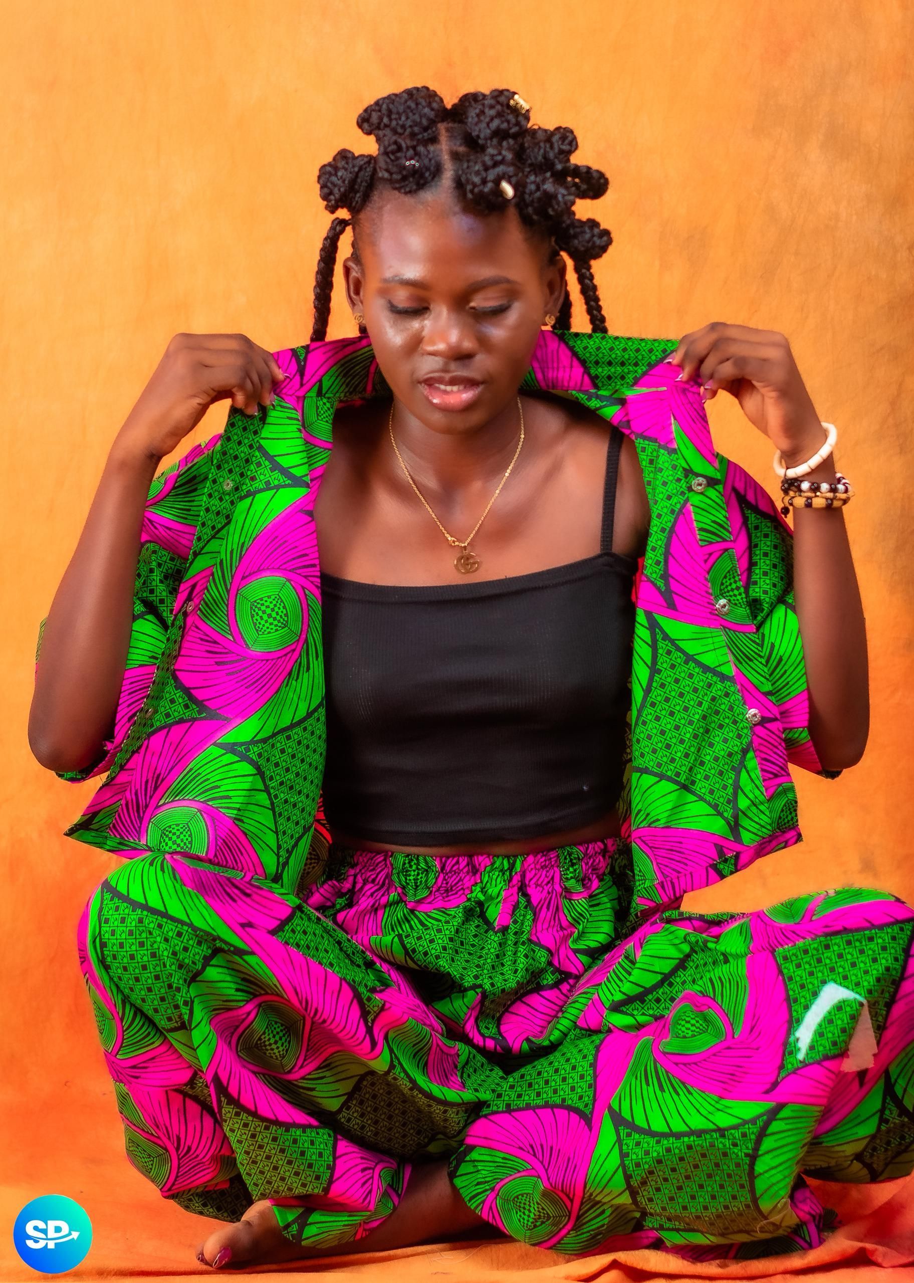 Une femme vêtue d'une tenue aux couleurs vives et à motifs est assise en tailleur. Elle ajuste sa veste.