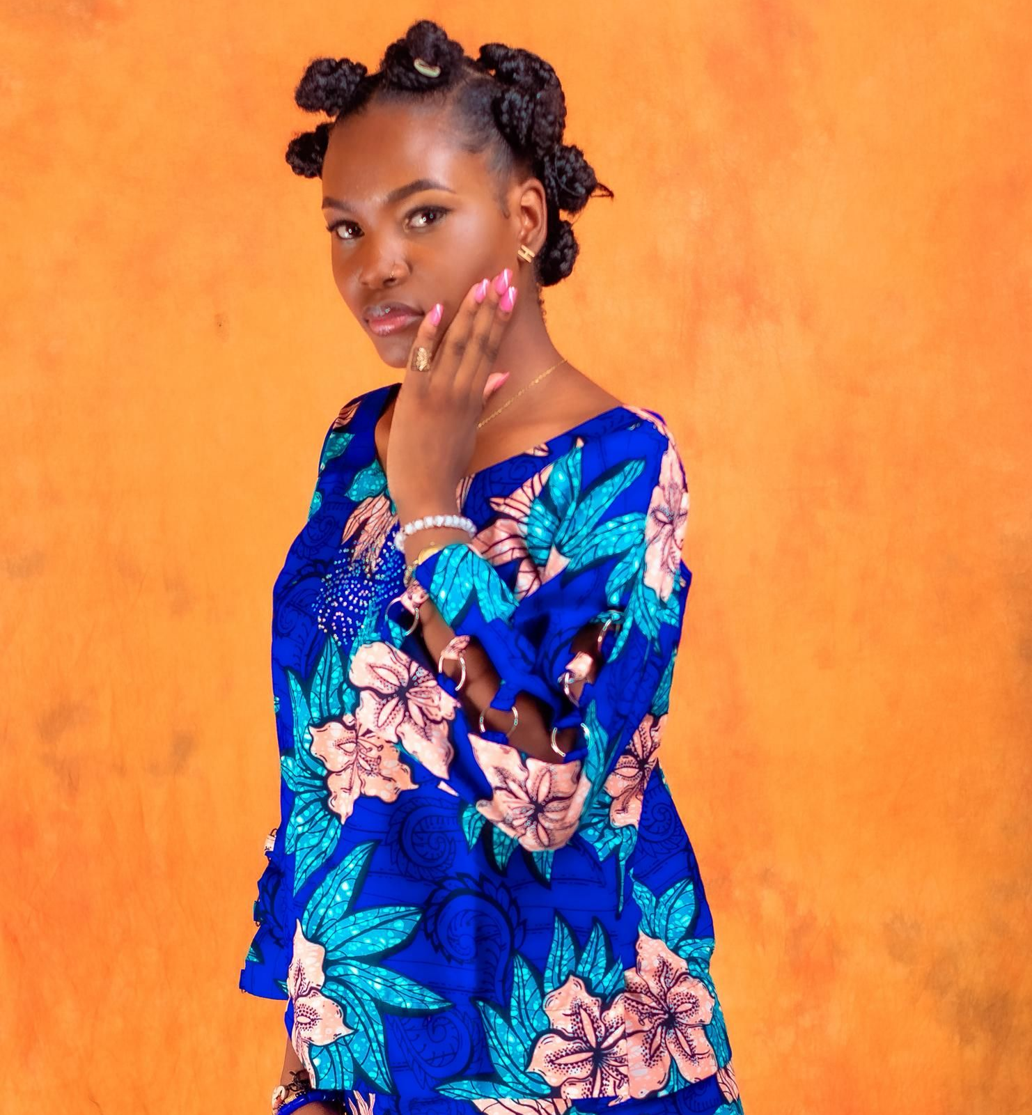 Femme en haut bleu à fleurs, coiffure tressée, posant devant un fond orange.