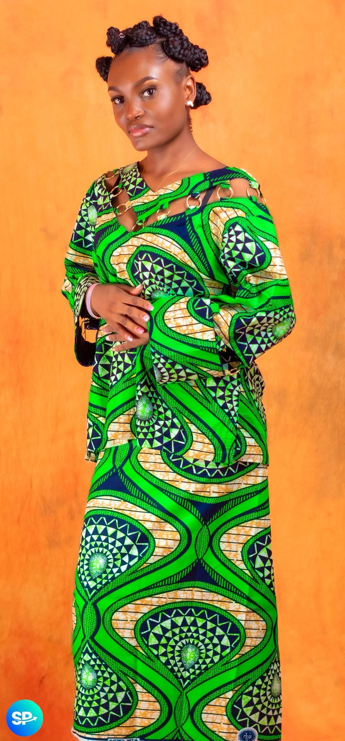 Une femme en robe verte à motifs pose devant un fond orange.