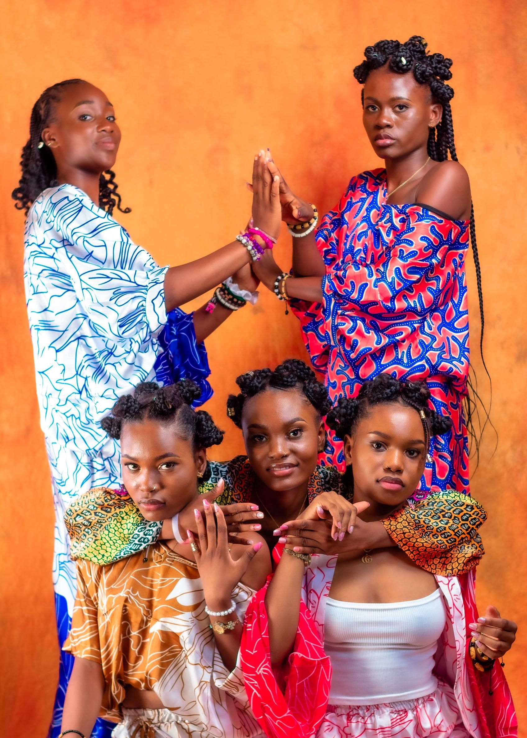 Cinq personnes vêtues de robes et de hauts à motifs colorés, bras dessus bras dessous, mains qui se touchent, sur fond orange.