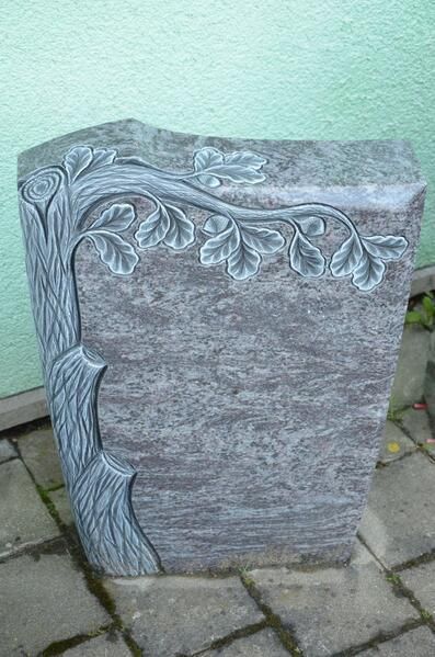 Auf einem gepflasterten Gehweg steht ein Grabstein mit einem eingemeißelten Baum.