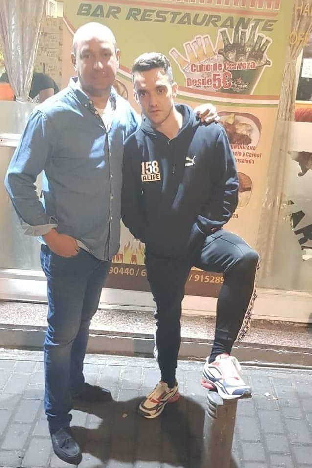 Dos hombres posando fuera de un bar. Uno con sudadera con capucha y el otro con camisa, ambos sonriendo.