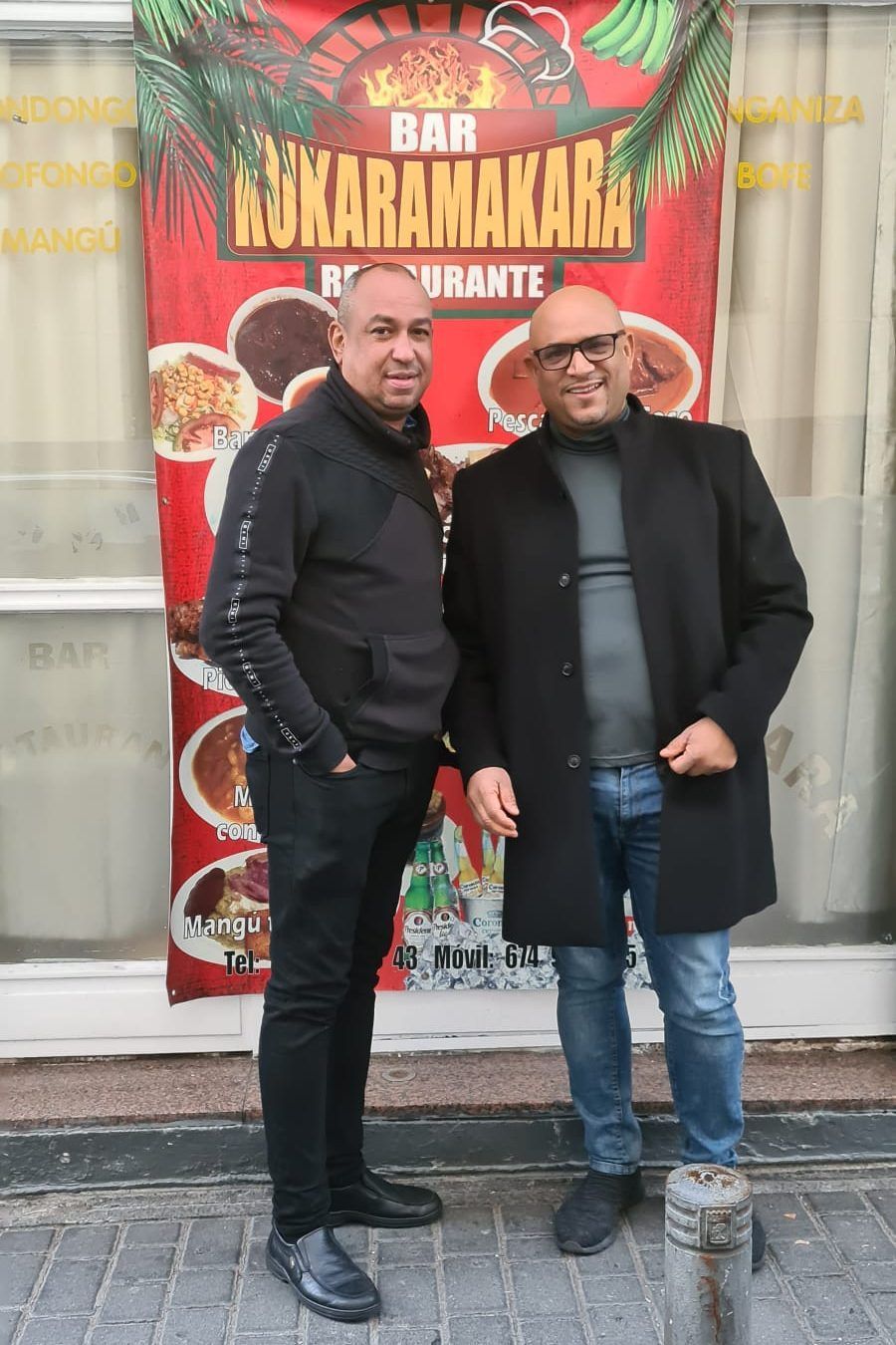 Dos hombres están frente al cartel de un restaurante. Uno lleva un suéter negro y el otro un abrigo negro.