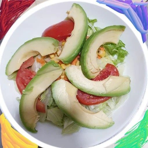 Ensalada con rodajas de aguacate, gajos de tomate y maíz sobre lechuga en un recipiente blanco.