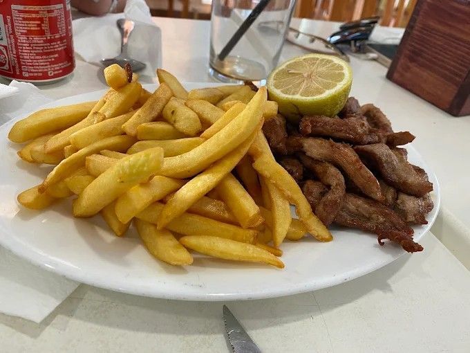 Plato de patatas fritas y carne con una rodaja de limón, junto a una bebida y un refresco.