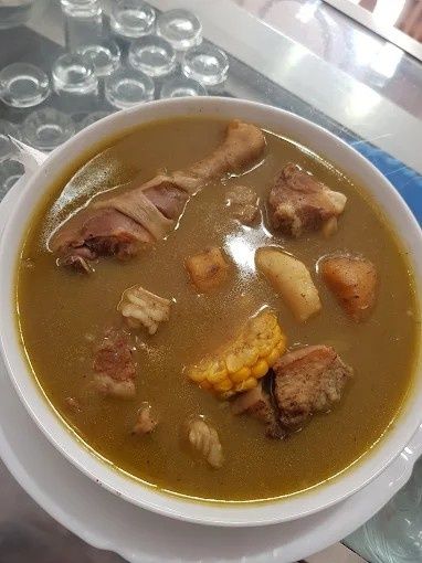 Sopa en recipiente blanco con muslo de pollo, trozos de carne, patatas y maíz.