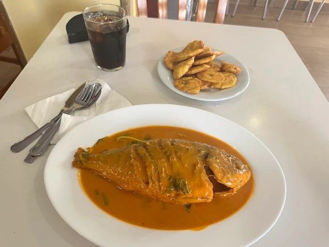 Pescado entero frito en salsa de naranja en plato blanco, chips de plátano, bebida, cubiertos.