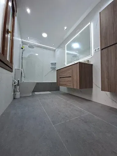 Baño moderno con suelo de baldosas grises, tocador flotante de madera, espejo grande y ducha a ras de suelo.