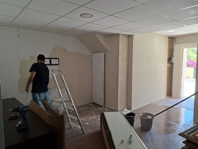 Persona pintando el interior de una habitación con escalera, herramientas y cubos de pintura en el suelo.