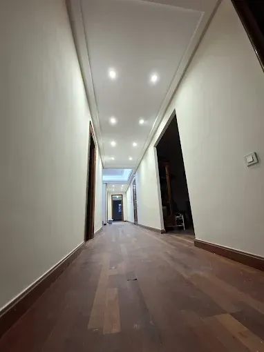 Largo pasillo vacío con paredes blancas, luces empotradas en el techo y suelos de madera oscura.