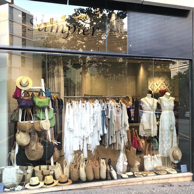 Escaparate de una boutique que exhibe ropa blanca de verano, bolsos de paja y sombreros dispuestos en percheros y estantes.