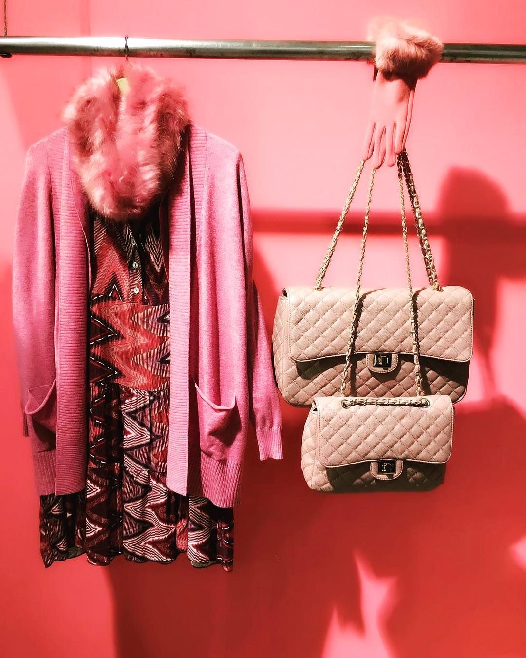 Un cárdigan rosa sobre un vestido estampado cuelga junto a dos bolsos acolchados con correa de cadena sobre un fondo rosa brillante.