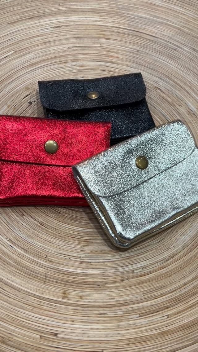 Tres pequeños monederos metálicos y brillantes de color rojo, negro y plateado, que descansan sobre una superficie circular tejida de color marrón claro.