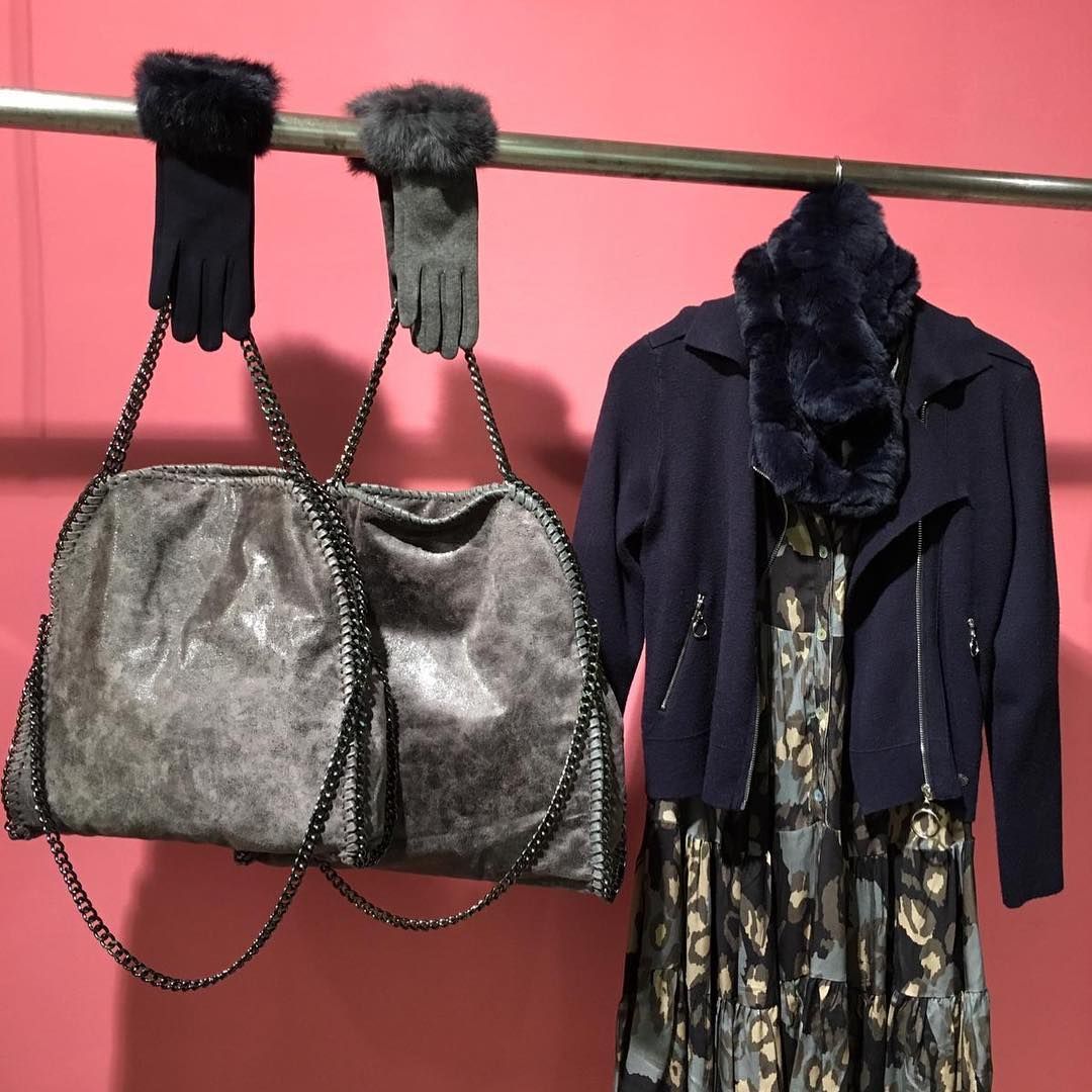 Dos bolsos metálicos cuelgan de unos guantes sujetos a una barra junto a una chaqueta y un vestido azul oscuro, contra una pared de color rosa vibrante.