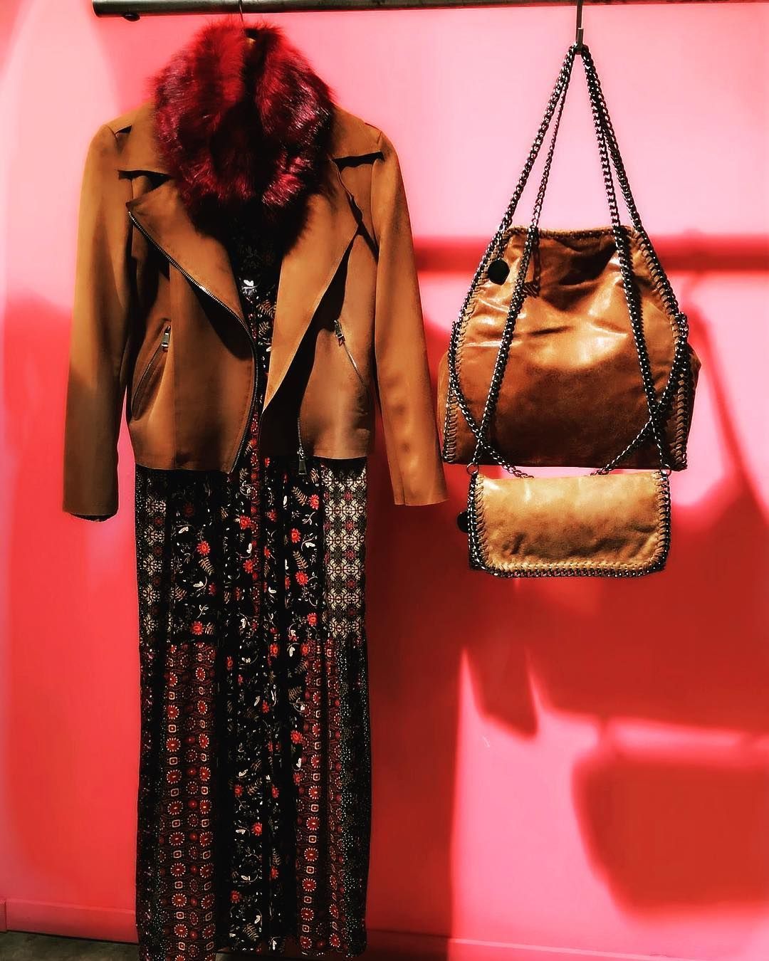 Una chaqueta color canela con cuello de piel roja, un vestido oscuro estampado floral y dos bolsos a juego color canela con correa de cadena colgados en un perchero.