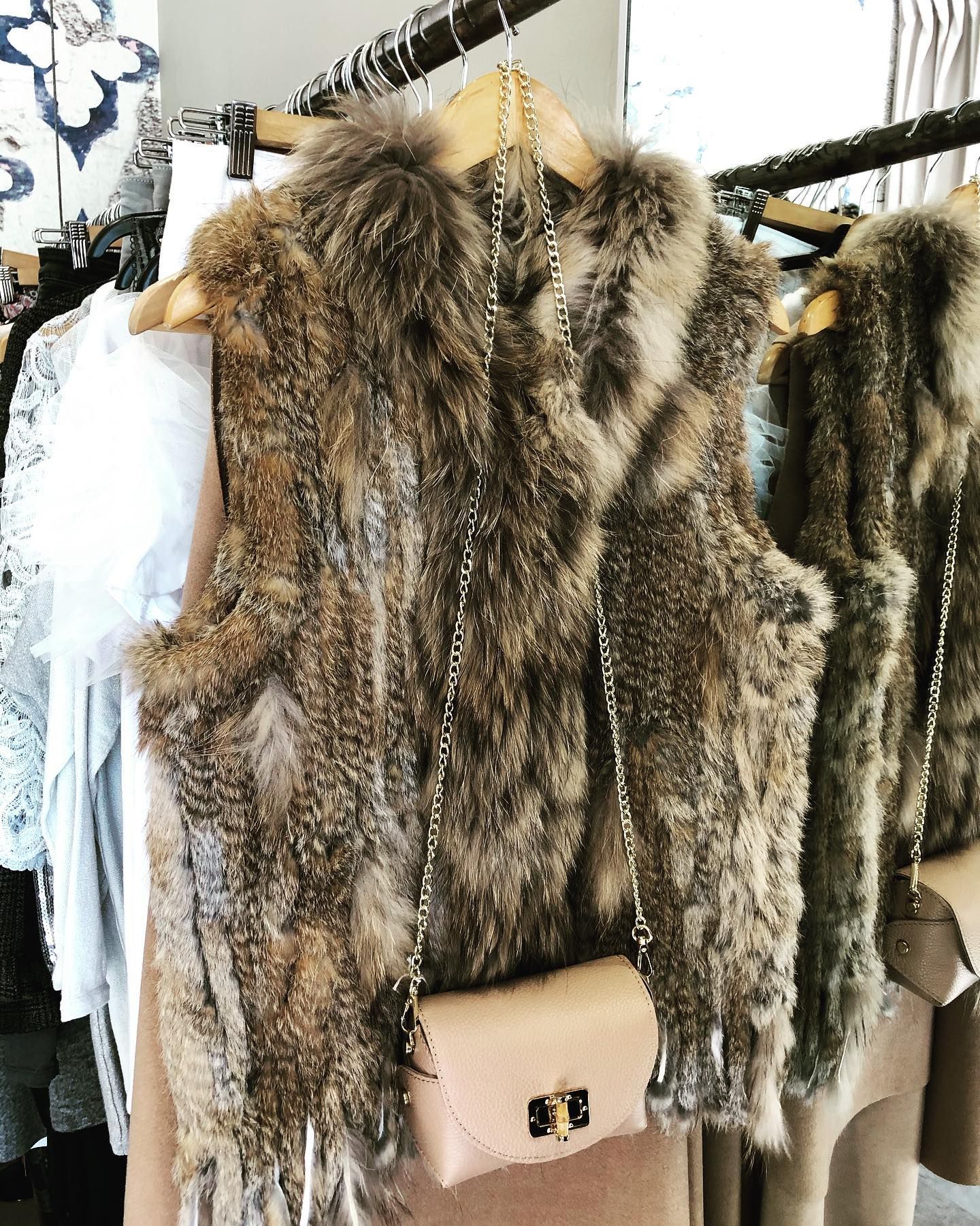 Un chaleco marrón de pelo sintético cuelga de un perchero, adornado con un pequeño bolso cruzado de color rosa claro con un broche dorado.