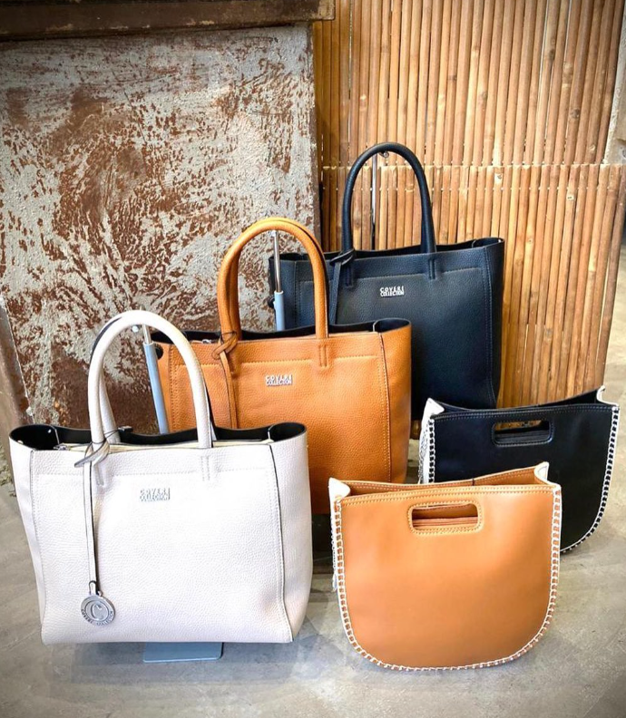 Una exhibición de cinco bolsos de mano en cuero blanco, marrón claro y negro, dispuestos sobre una superficie con fondo texturizado.