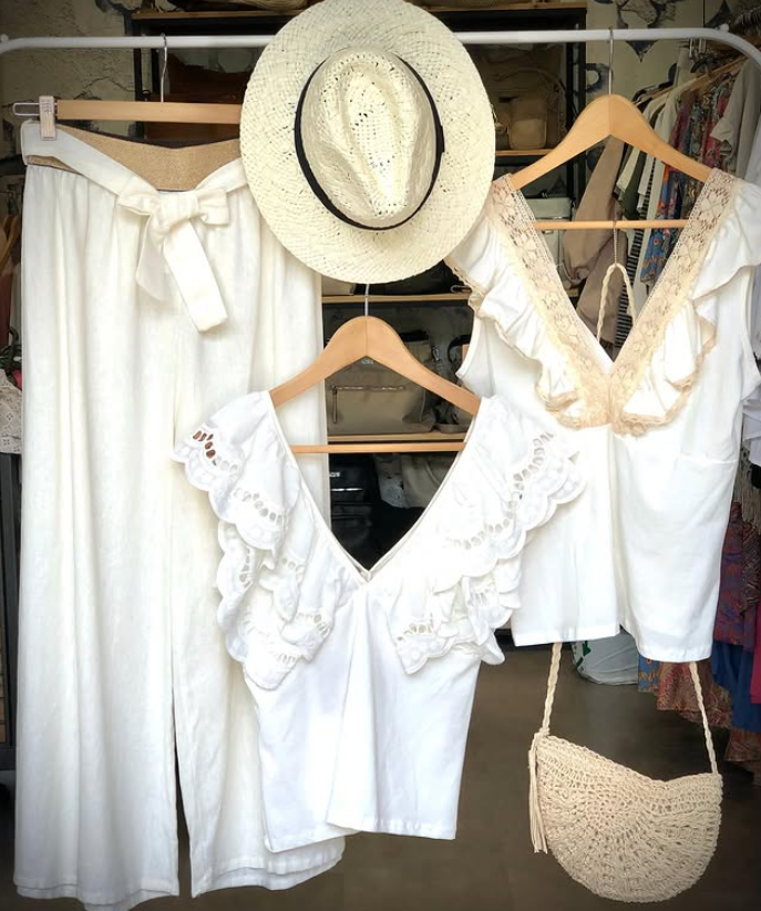 Pantalones blancos de pierna ancha, dos blusas blancas con volantes, un sombrero de paja y un bolso de mimbre colgado en un perchero.