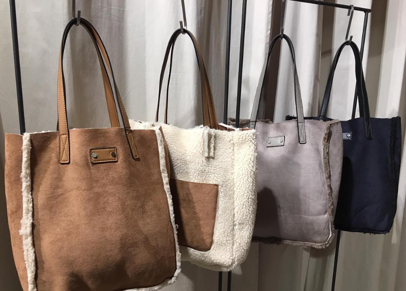 Cuatro bolsos de mano colgados en un perchero, con diseños que van desde ante color camel y borrego crema hasta modelos grises y negros.