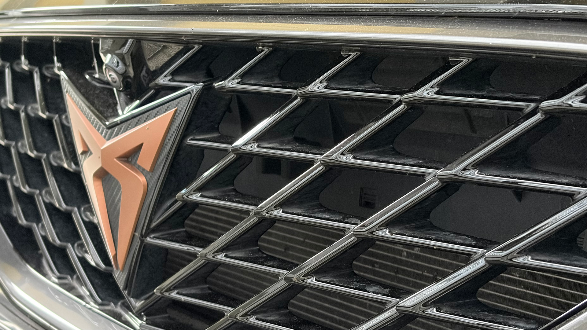 Nahaufnahme des kupferfarbenen Cupra-Logos auf einem dunklen, wabenförmigen Kühlergrill.