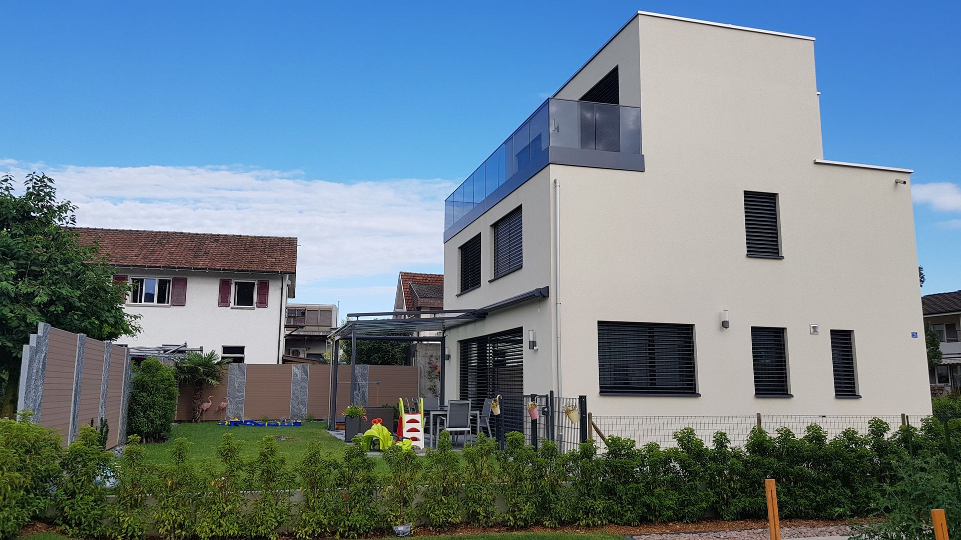 Modernes zweistöckiges beiges Haus mit verglaster oberer Etage und Balkon; grüner Rasen, blauer Himmel.