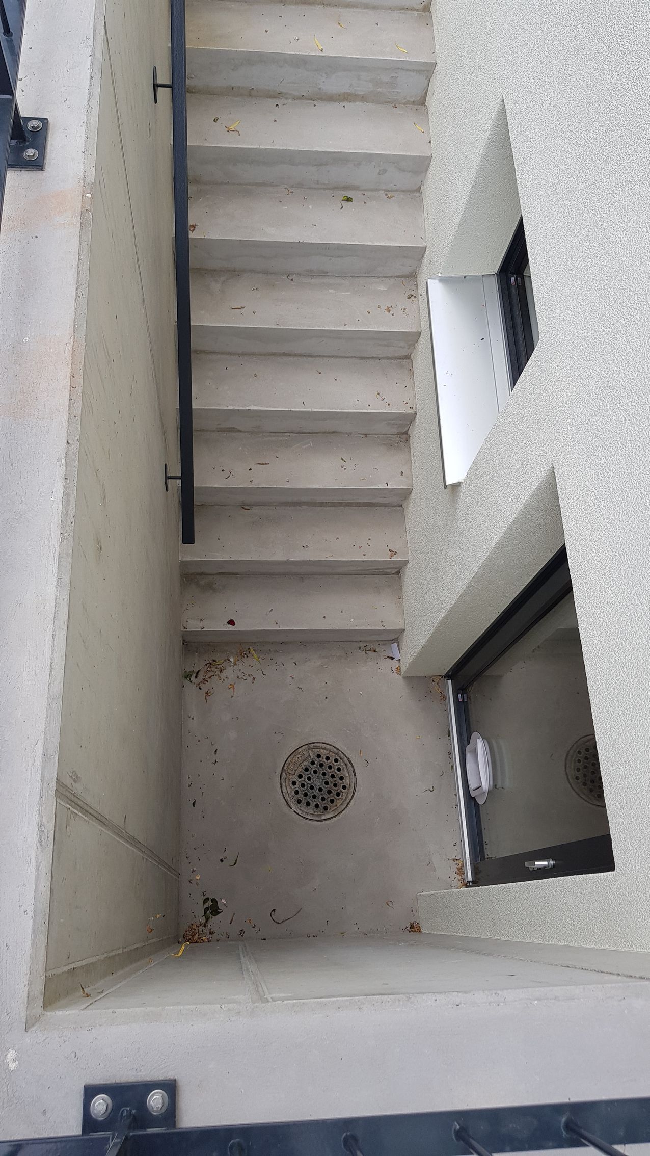 Betontreppe im Freien, die nach unten führt, mit einem schwarzen Geländer auf einer Seite. Strukturierte weiße Gebäudewand mit einem Fenster.
