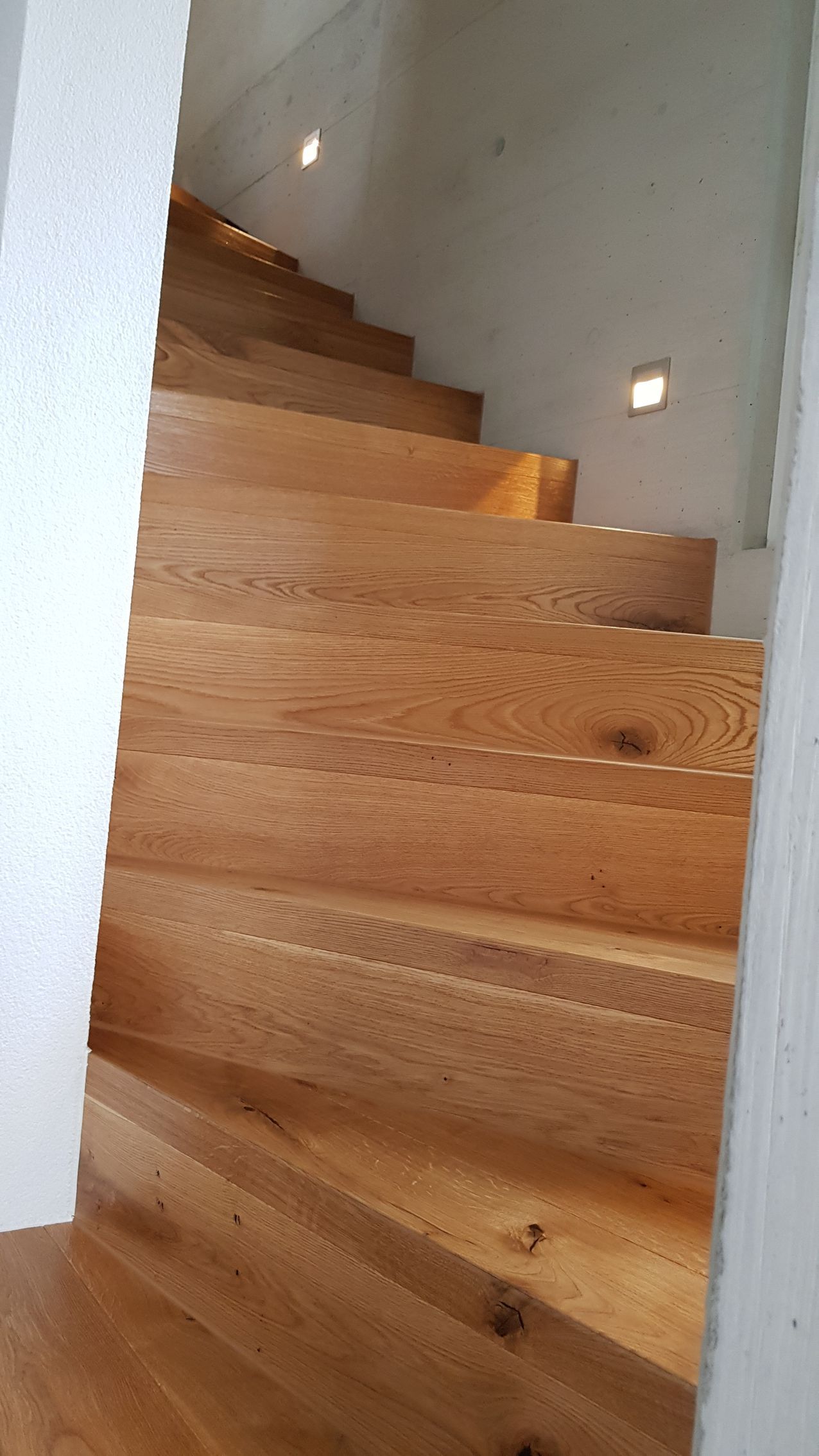 Hölzerne Wendeltreppe mit Einbauleuchten in einem Flur mit weißen Wänden.